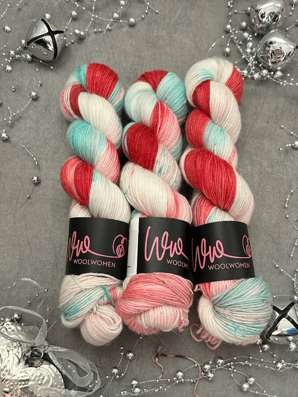 Three skeins of WoolWomen Alpaca Sock - yhden padan ihmeet yarn in red, mint green, and white are displayed on a gray surface with silver beads and festive decor. Each skein is labeled "Woolwomen.