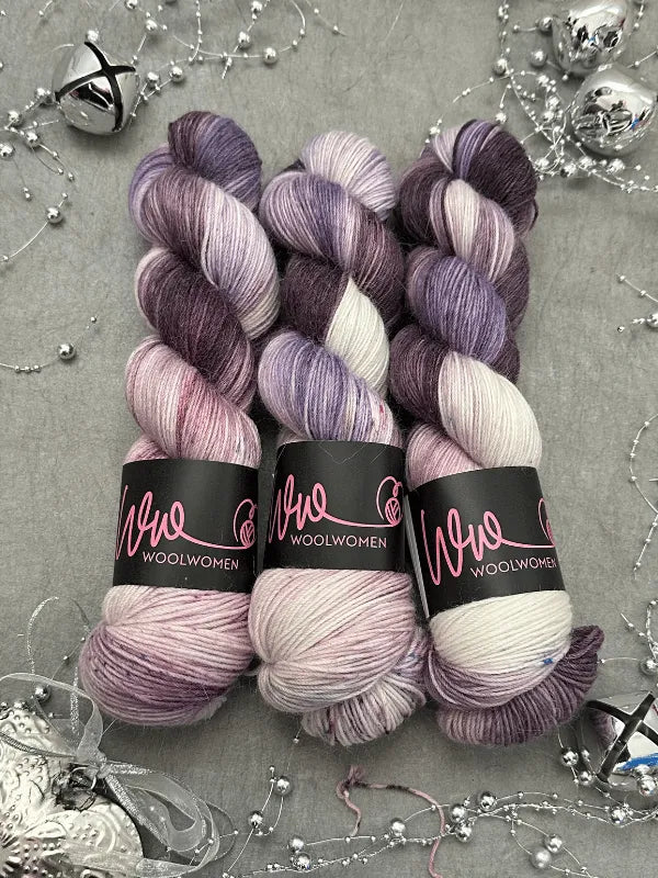 Three skeins of WoolWomen Alpaca Sock - yhden padan ihmeet yarn in variegated purple, lavender, and white are displayed on a gray surface, surrounded by silver beads, tinsel, and jingle bells.