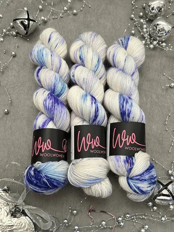 Three skeins of WoolWomen Alpaca Sock - yhden padan ihmeet in white with blue and purple speckles are arranged on a gray surface, surrounded by silver bells, beads, and decorative ornaments.