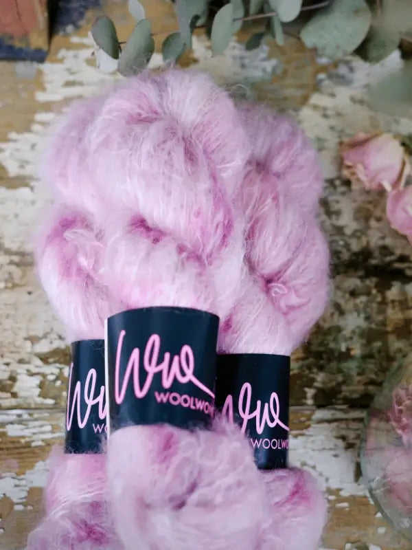 Three skeins of soft, fluffy pink WoolWomen Fluff Mohair yarn are stacked on a rustic wooden surface, with dried flowers and greenery in the background.