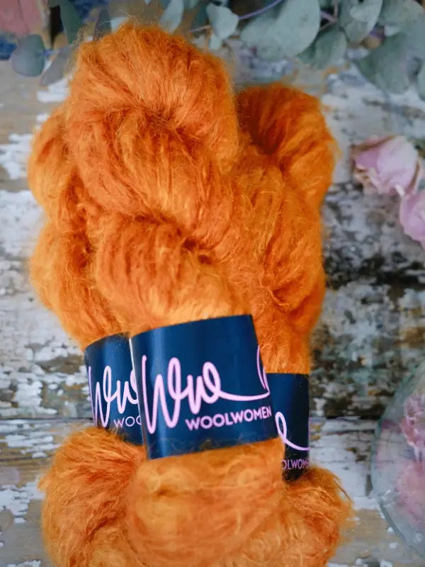 Two skeins of bright orange WoolWomen Fluff Mohair rest on a rustic wooden surface, surrounded by dried leaves and petals. This käsinvärjätty mohairlanka brings vibrant color and softness to any project.