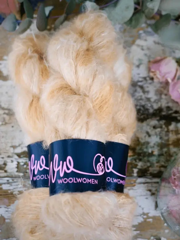 A skein of light tan WoolWomen Fluff Mohair with a black label in pink script rests on a rustic, weathered surface scattered with leaves and flowers.