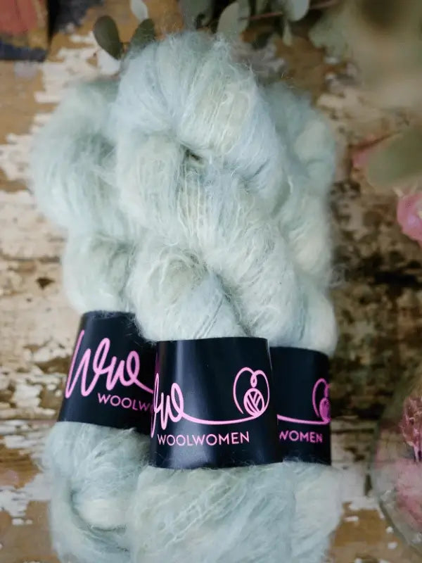 Two skeins of pale mint green WoolWomen Fluff Mohair, with black labels and pink text, rest on rustic wood—ideal for your next Fussa neuletakki neuleohje project.