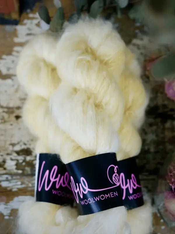Two skeins of WoolWomen Fluff Mohair, soft and pale yellow with black labels in pink script, rest on a rustic wooden surface. This käsinvärjätty mohairlanka is ideal for cozy handmade projects.