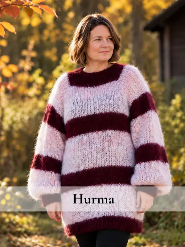 A woman stands outdoors in autumn, wearing a long, cozy sweater with wide maroon and light pink stripes from the Hurma neulesetti – WoolWomen Fluff Mohair. Colorful fall foliage surrounds her, with “Hurma” overlaid on the image.