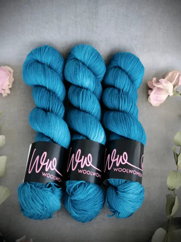 Three skeins of vibrant teal WoolWomen Merino Sock yarn are arranged side by side on a gray surface, accented with soft pink flowers and green foliage at the corners.