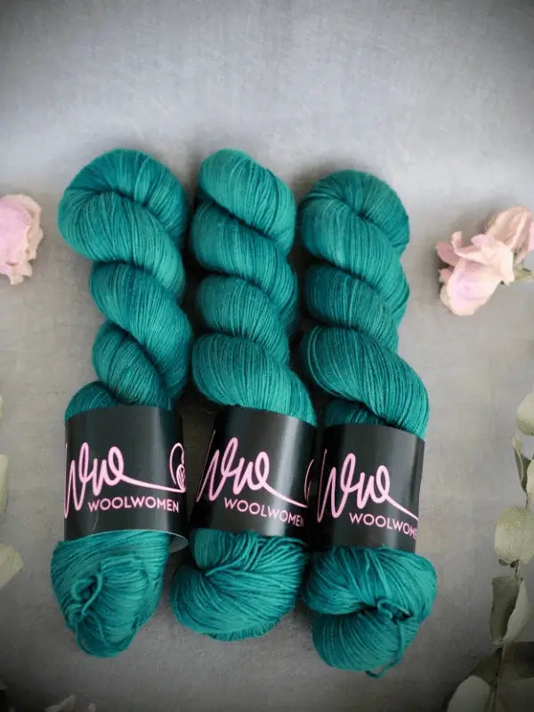 Three skeins of WoolWomen Merino Sock yarn in teal, käsinvärjätty merinovilla, are arranged side by side on a gray surface, with light pink flowers around the edges.