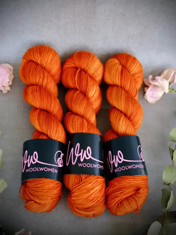 Three skeins of bright orange WoolWomen Merino Sock yarn with black Woolwomen labels are arranged side by side on a gray surface, surrounded by several pink and green flowers.