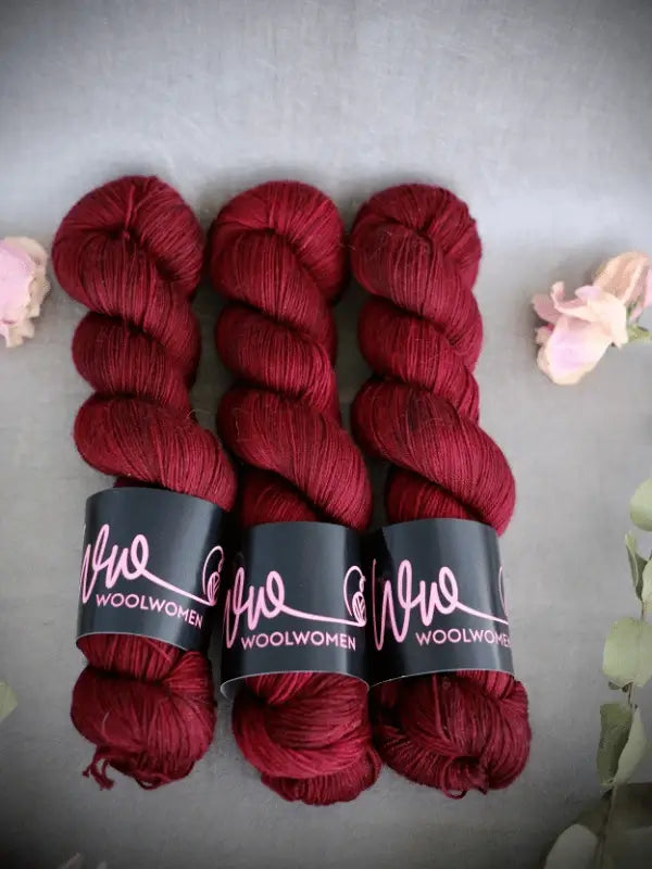 Three skeins of deep red WoolWomen Merino Sock yarn are arranged side by side on a gray surface, with soft pink flowers and green leaves around them.