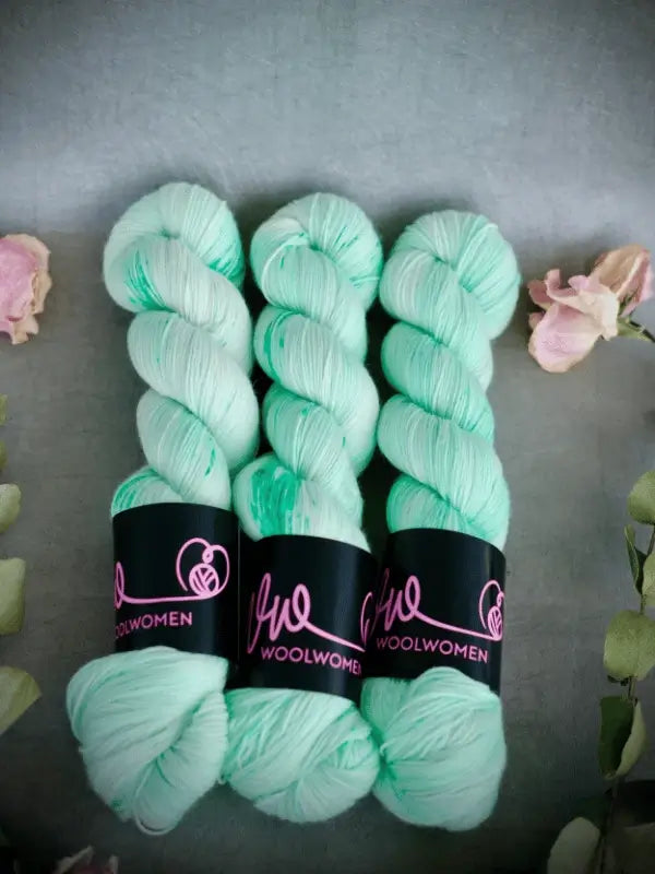 Three skeins of mint green WoolWomen Merino Sock yarn, accented with darker green speckles and wrapped in black labels with pink text, are arranged side by side on a gray surface adorned with floral decorations.