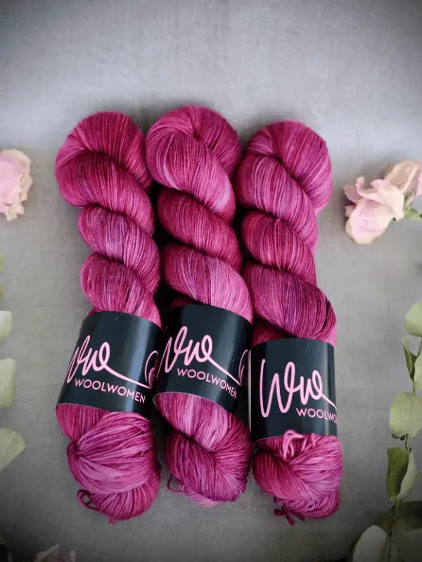 Three skeins of bright magenta WoolWomen Merino Sock yarn with black labels are neatly arranged on a gray surface, surrounded by soft pink and green floral accents.