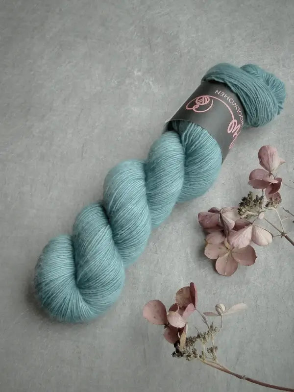 A skein of WoolWomen Merino Single in light blue lies on a gray surface beside a small dried flower arrangement with pale pink and purple petals.