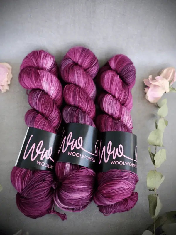 Three skeins of magenta and purple variegated WoolWomen Merino Sock yarn are displayed side by side on a gray surface, surrounded by pink flowers and green leaves.