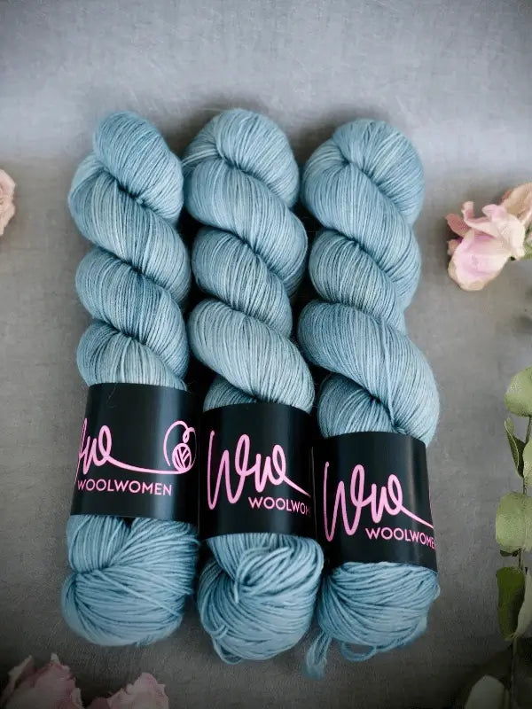 Three skeins of light blue WoolWomen Merino Sock yarn with black labels and pink text are arranged side by side on a gray surface, surrounded by soft pink flowers.