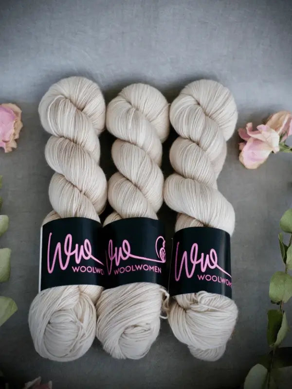 Three skeins of WoolWomen Merino Sock in cream are arranged side by side on a gray surface, with light pink flowers and green leaves placed around them.