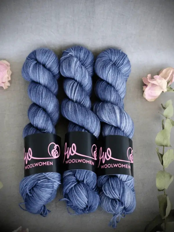 Three skeins of blue WoolWomen Merino Sock yarn are arranged side by side on a gray surface, with pink and green dried flowers placed around them.