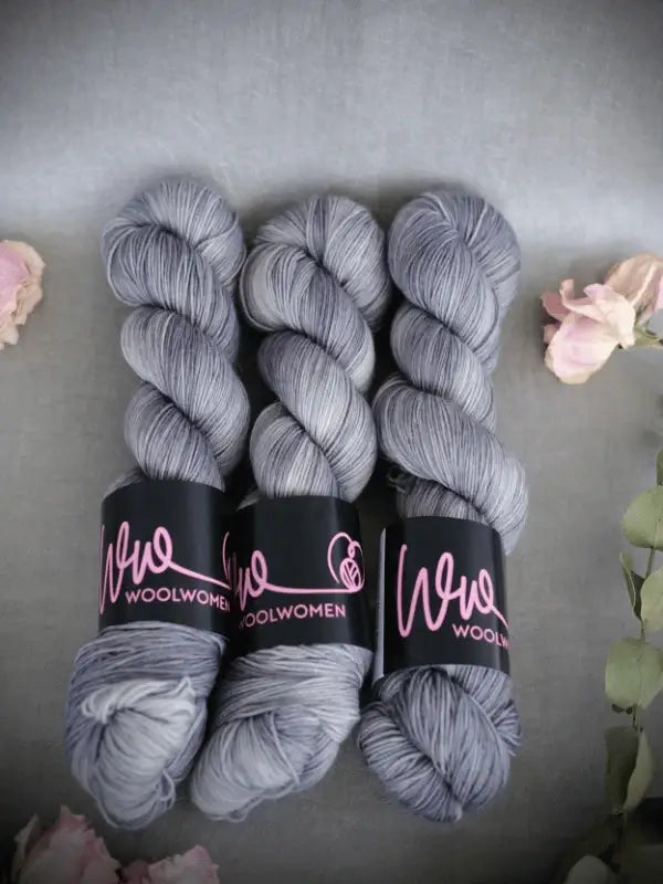 Three skeins of light gray WoolWomen Merino Sock yarn with black and pink tags are arranged side by side on a gray surface, surrounded by pink flowers and green leaves—premium superwash merinovilla käsinvärjätty lanka.