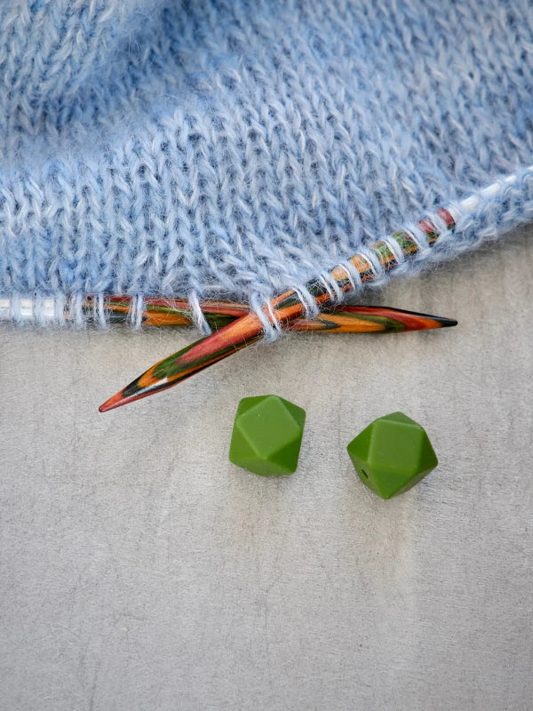 Two colorful Woolwomen Puikkostopparit wooden knitting needles with light blue yarn and two green faceted beads rest on a gray surface.