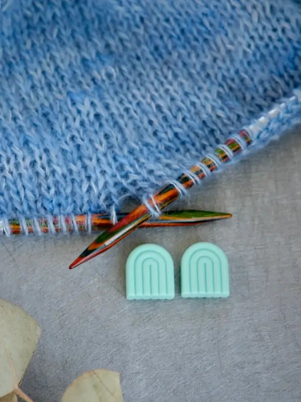A close-up of blue knitted fabric on colorful knitting needles, with two mint green Woolwomen Puikkostopparit arch-shaped accessories lying on a gray surface below.