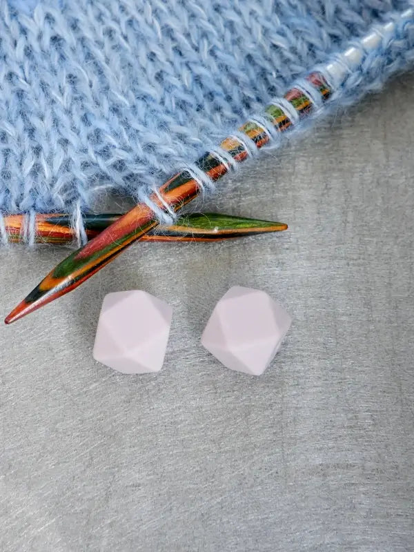 A close-up of two pink, faceted Woolwomen Puikkostopparit on a gray surface beside colorful knitting needles and light blue knitted fabric.