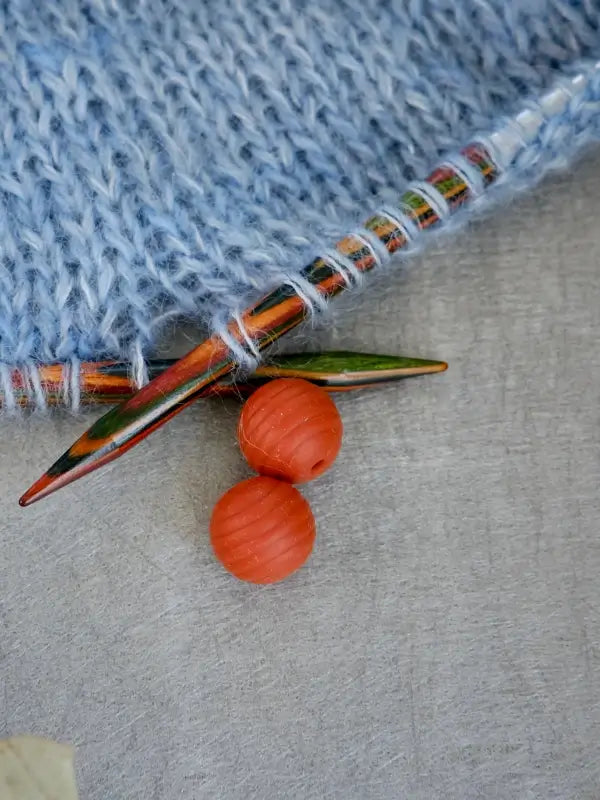 Close-up of a light blue knitted fabric on colorful knitting needles, with two round orange Woolwomen Puikkostopparit resting nearby on a light gray surface.