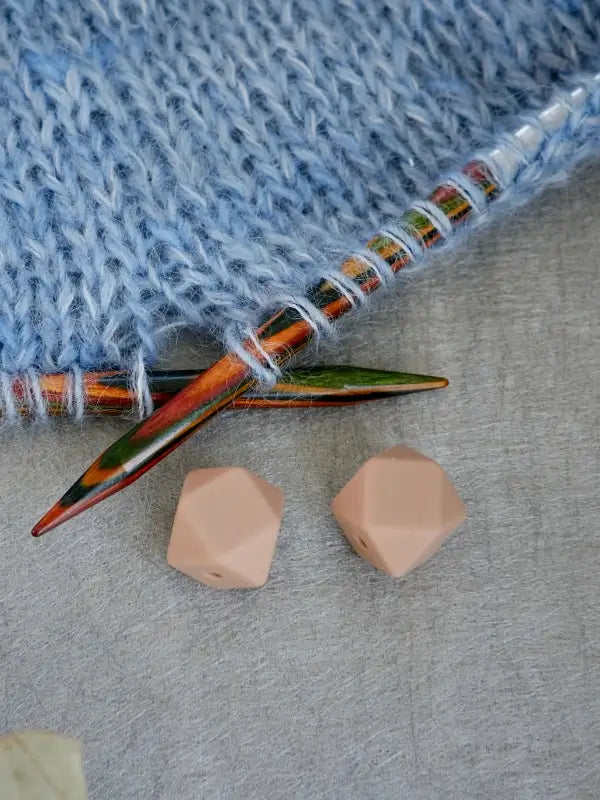 A close-up of blue knitted fabric on colorful wooden needles, with two light brown Woolwomen Puikkostopparit geometric beads placed nearby on a gray surface.