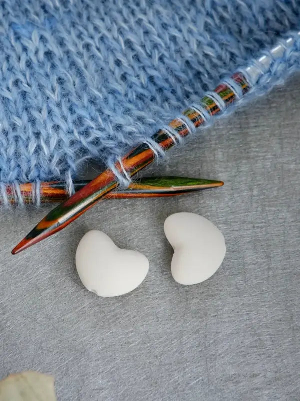 A pair of colorful knitting needles rests on blue knitted fabric, with two Woolwomen Puikkostopparit—white, heart-shaped needle stoppers—placed on a gray surface nearby.