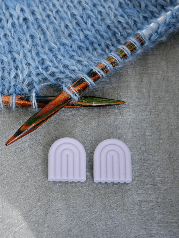 A pair of wooden knitting needles with blue yarn beside two Woolwomen Puikkostopparit lavender silicone needle point protectors on a gray surface.