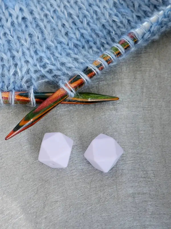Two vibrant Woolwomen Puikkostopparit knitting needles with blue yarn rest beside two white, faceted beads on a gray surface.