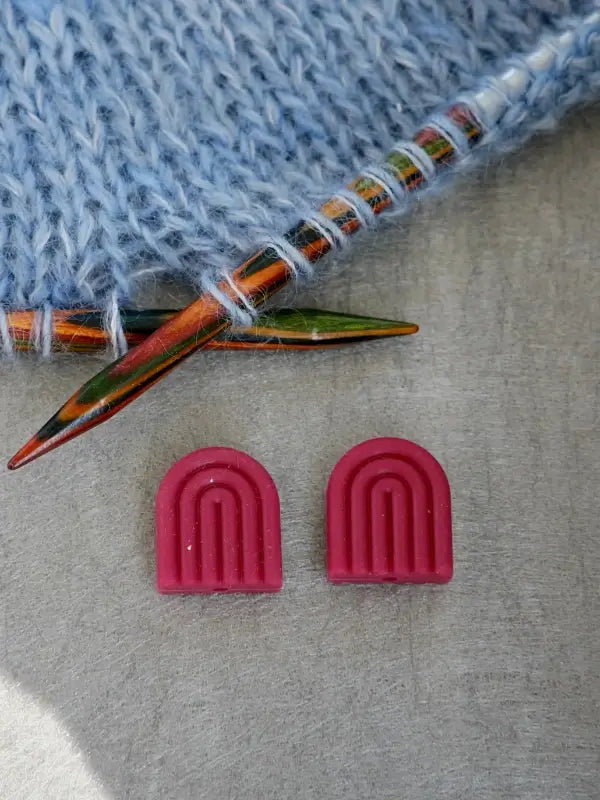 A close-up of two colorful knitting needles in blue yarn, with two pink Woolwomen Puikkostopparit rainbow-shaped stitch markers on a gray surface below.