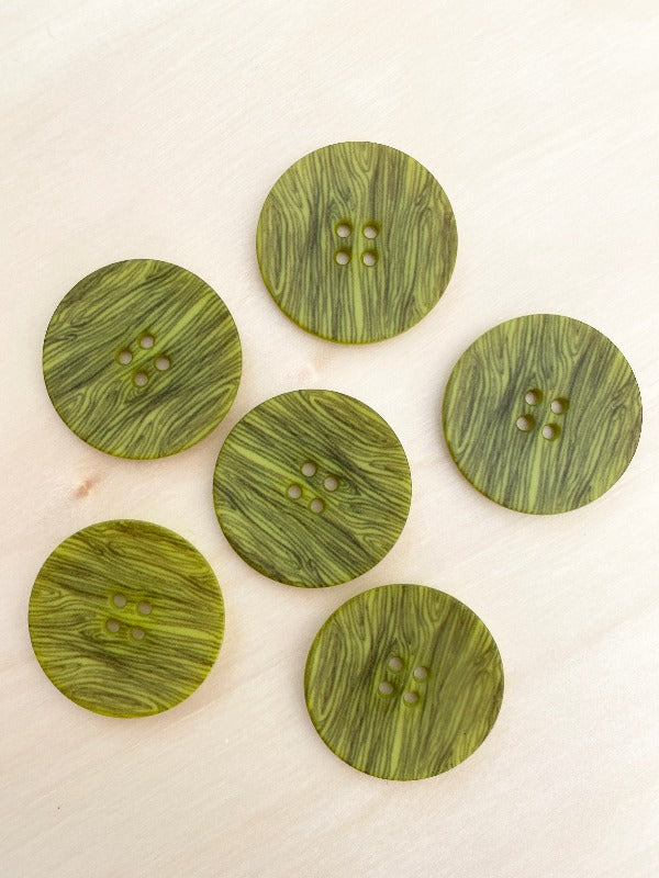 Six green Muovinappi 23mm buttons with a woodgrain pattern and four holes are displayed on a light surface. These lightweight buttons are available in various colors.