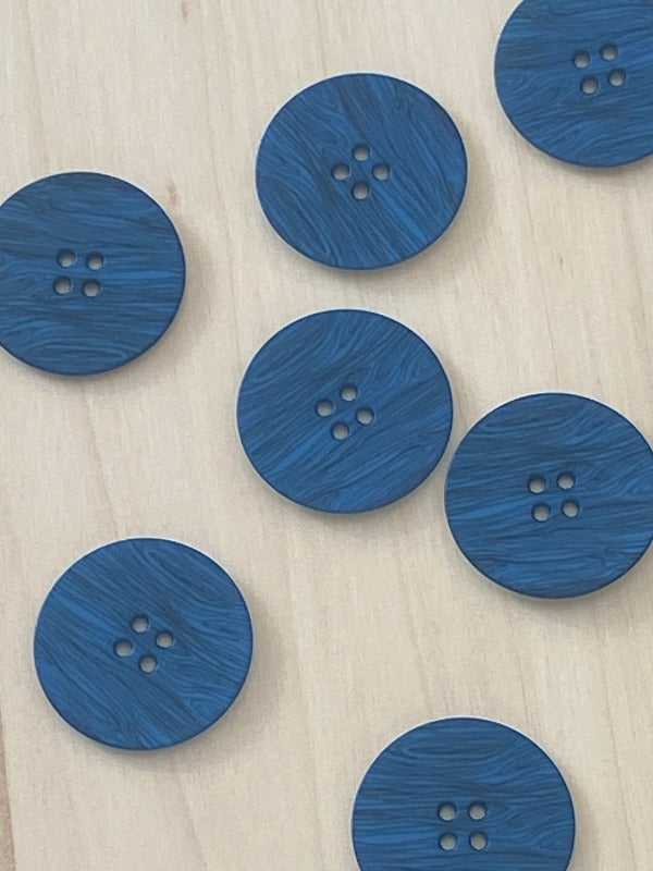 Seven Muovinappi 23mm round blue buttons with a woodgrain texture and four holes are scattered on a light wooden surface. These lightweight (kevyt) buttons are available eri väreissä to match your style.